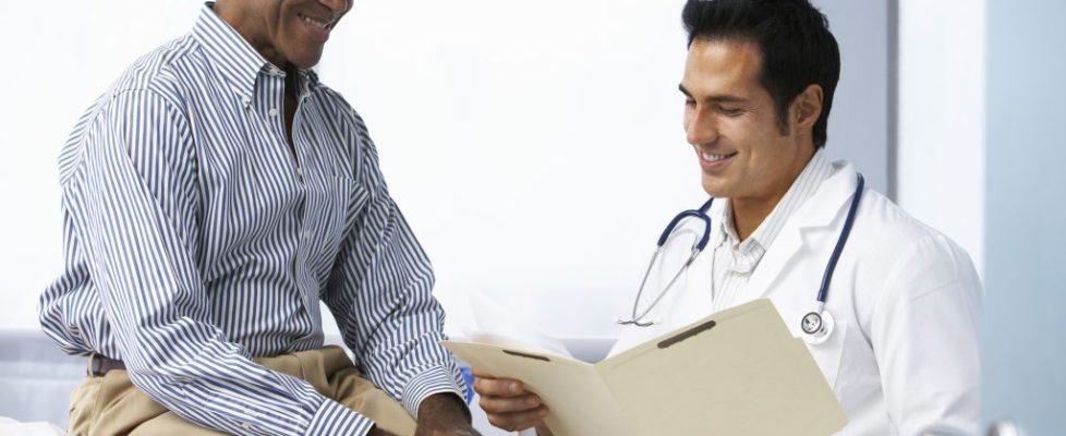Doctor In Surgery With Male Patient Reading Notes Doctor In Surgery With Male Patient Reading Notes